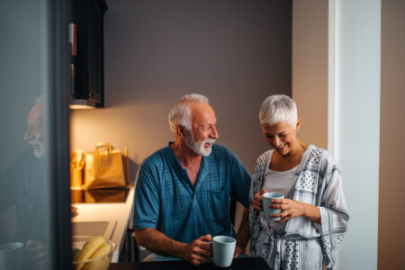 An older man and woman stand in a kitchen, smiling and holding coffee mugs