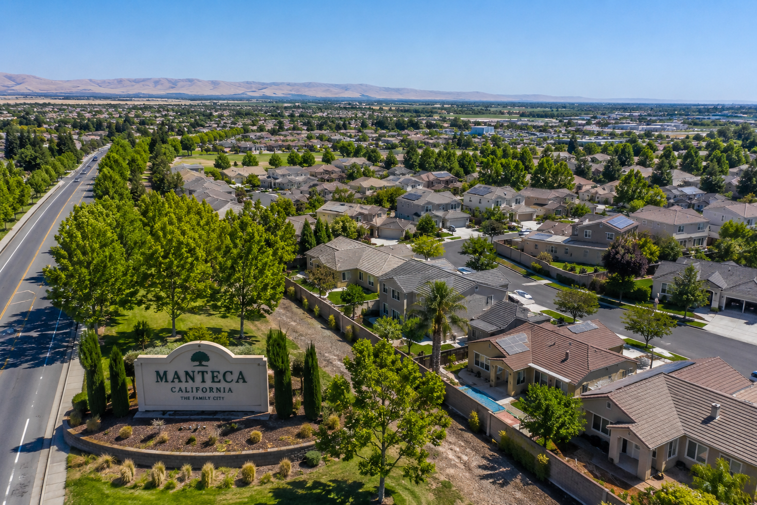 Aerial view of a residential neighborhood in Manteca, California, featuring tree-lined streets, single-family homes, and the Manteca city welcome sign on a clear sunny day.