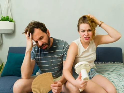 Tired man and woman waving handheld fan and newspaper, trying to cool off as room is hot even with ac running and the rest of the house is cool.